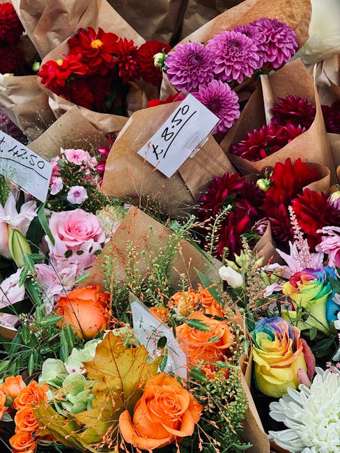 A vibrant assortment of fresh flowers, including red dahlias, purple chrysanthemums, pink roses, orange roses, and multicoloured rainbow roses, arranged in paper wrapping for flower delivery or giftin