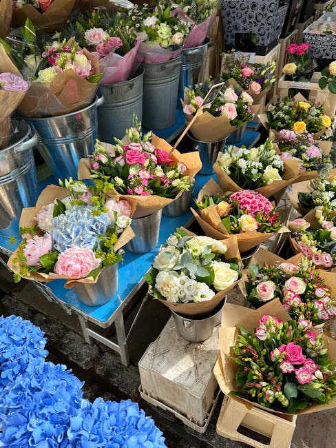 An outdoor display of various freshly arranged flower bouquets and floral gifts set on tables and in metal buckets, featuring pastel pink, white, and blue flowers including roses, hydrangeas, and othe