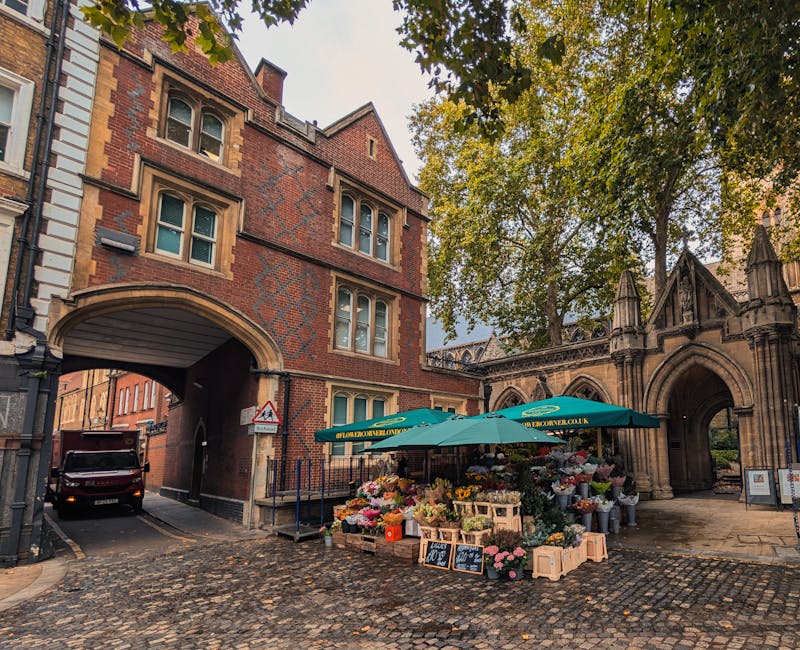 A vibrant flower stall situated on a cobblestone street in front of an old brick and stone building with Gothic architectural features and an arched gateway. The stall, shaded by a teal umbrella with 