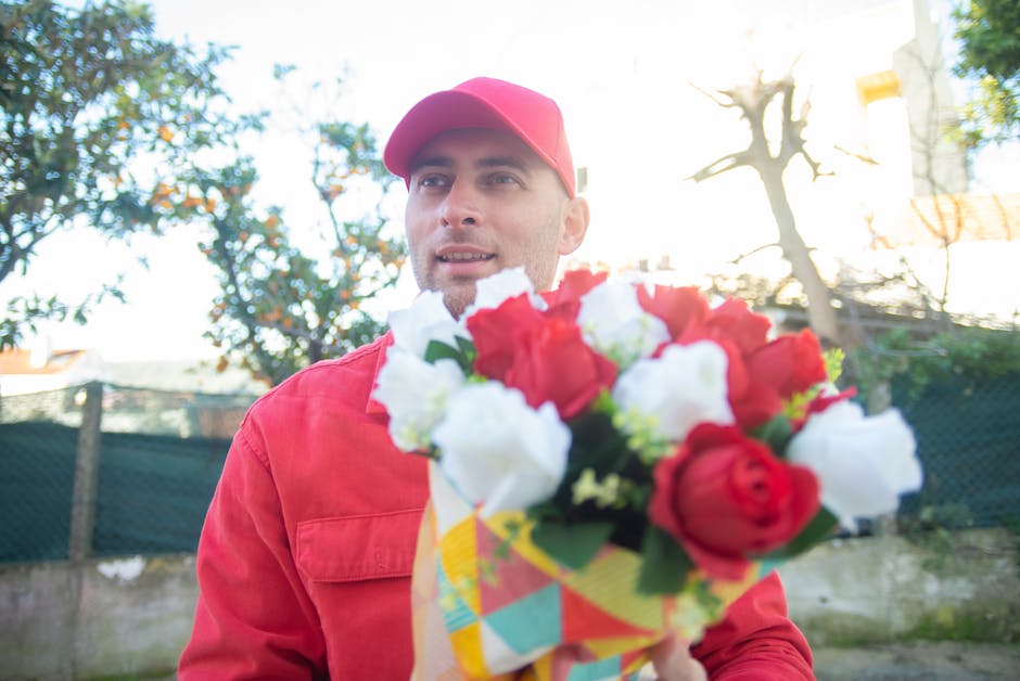 A male flower delivery courier, dressed in a red uniform and cap, stands outdoors in natural daylight holding a vibrant bouquet of fresh flowers, including red and white blooms with green stems and le