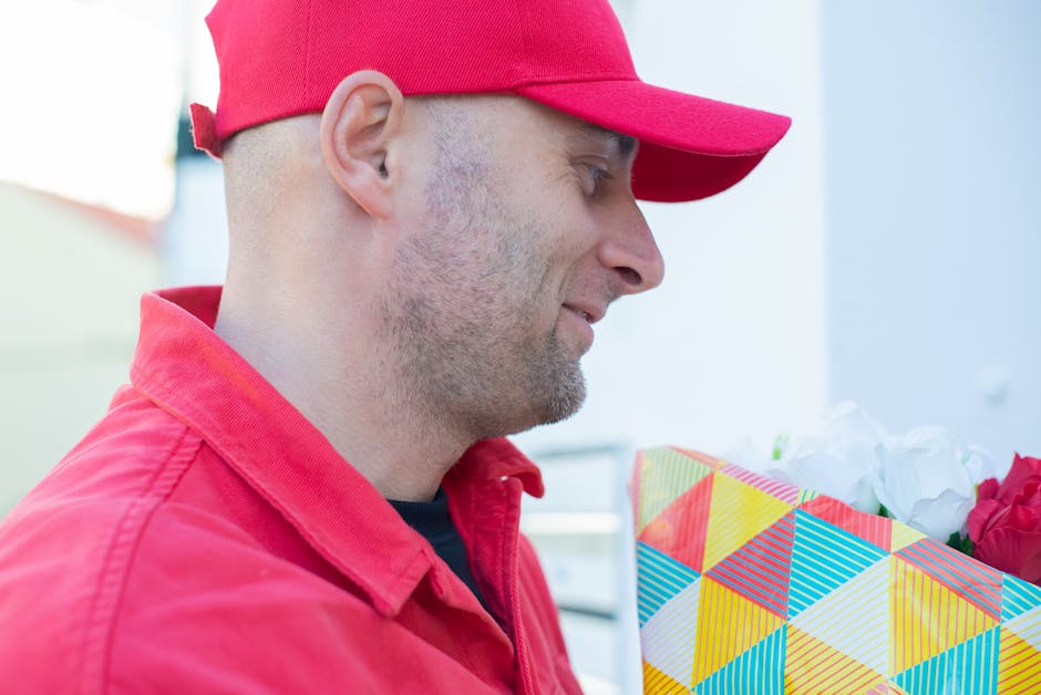 A man wearing a red baseball cap and matching red jacket is seen in profile, smiling softly while holding a colorful, geometric-patterned gift bag filled with fresh flowers. The bouquet inside feature