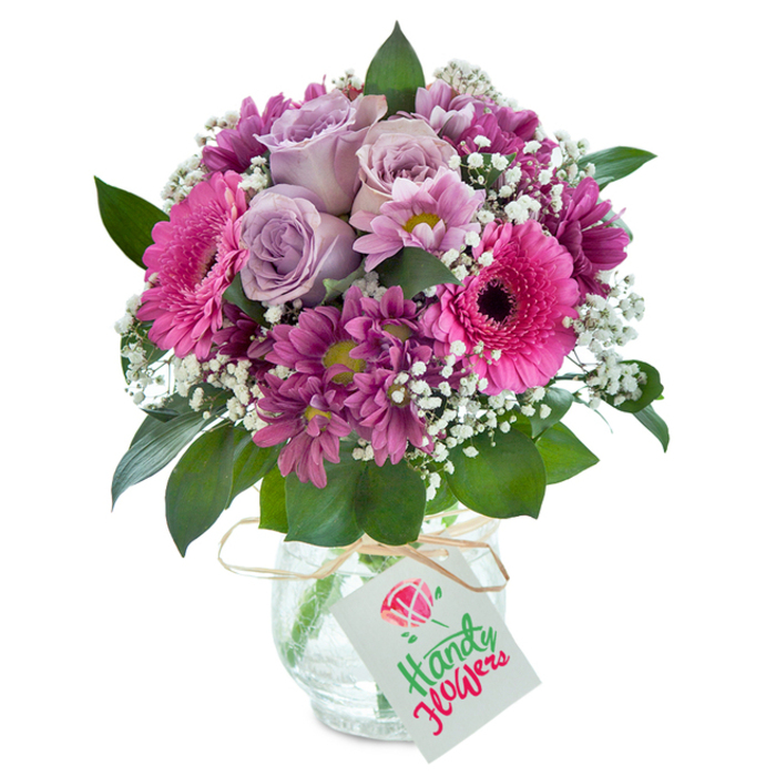 Taking centre stage against a clean white background, this Colliers Wood bouquet is a celebration of vibrant artistry and local charm. The arrangement is anchored by soft lavender roses, their petals unfolding in layered elegance, carrying hints of gentle perfume like those from gardens nestled beside Wandle Park. Large, spirited Gerbera daisies in brilliant fuchsia-pink stand tall, their signature dark centres creating lively contrast. Magenta-hued chrysanthemums-some with hints of green at their centres-cluster tightly, bringing depth and a sense of abundance that echoes Colliers Wood's community feel. Delicate baby's breath is tucked in throughout, lending an airy, almost magical brightness that lifts the whole design. Dense, dark green leaves frame the bouquet, creating a lush foundation that displays the colours to their fullest. Everything is gathered neatly into a clear, crackled glass vase that diffuses the light and adds a touch of rustic flair. A length of natural raffia secures a simple white tag at the vase's neck, waiting for a meaningful handwritten note. This arrangement spotlights the skill and care of local floristry, making it perfect for birthdays, farewells, or a heartfelt thank-you-delivered with pride by Colliers Wood Florist to your door.